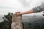 Anonymer Wanderer, der seine Hand auf einem rauen Stein vor der unscharfen Landschaft eines bewaldeten Hochlands hält