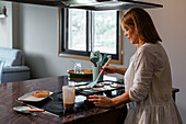 Side view of adult woman in white shirt standing at counter and flipping pancakes with spatula while making breakfast