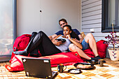 Positives junges gemischtrassiges Paar ruht sich auf einer Couch aus und schaut einen Film auf einem Laptop auf dem Balkon