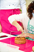 Crop unrecognizable female with professional equipment cutting fresh baked cake on parts in bakery
