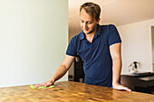 Young positive male wearing casual navy blue polo shirt wiping wooden dining table with yellow cloth