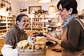 Side view of positive female seller reaching out hand with terminal to happy senior woman doing payment with credit card in eco friendly store
