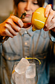 Soft focus of crop female barkeeper taking off lemon zest into glass with ice cubes while making cocktail in bar