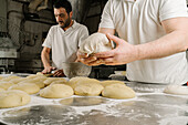 Cropped mature bearded ethnic bakers forming bread from dough at table with flour and bowl in bakery