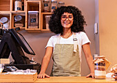 Young smiling African American female seller in apron and glasses looking at camera standing near of modern cash register during work in zero waste grocery store