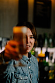 Female bartender looking at camera while serving wineglass with gin and tonic cocktail decorated with orange zest on wooden counter in bar