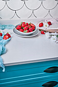 From above bowl of ripe strawberries placed on counter drawer near fresh white flowers and opened book in daytime at home
