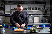 Bearded male chef cutting fresh artichokes while preparing delectable dish on table in kitchen of contemporary restaurant