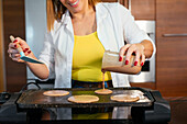 Cropped unrecognizable cheerful adult woman pouring batter on hot electric griddle while cooking pancakes at counter in modern kitchen