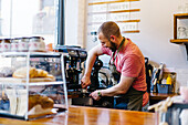 Side view of male barista in apron with portafilter brewing coffee using coffee machine while standing near counter in coffee house