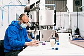 Male quality control specialist in sterile mask studying alcoholic liquid at table against professional equipment in factory