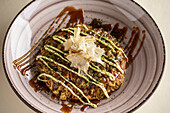 From above of tasty traditional Japanese okonomiyaki dish with sauce topped with bonito flakes served in ceramic bowl on light background