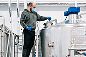 Side view of male engineer in face mask examining stainless steel tank with hoses in brewery with wet floor during pandemic