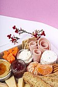 From above brunch box with assorted sliced meats various types of cheese and crispbreads arranged near ripe cup kiwi sweet strawberries and peeled mandarin near jam in glass jar on colorful background