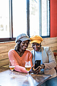 African American friendly females taking self shot on smartphone while sitting at table with glasses of beer in bar and enjoying weekend