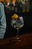 Crop unrecognizable bartender serving wineglass with gin and tonic cocktail decorated with orange zest on wooden counter in bar