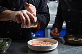 Anonymer Mann, der Paprika über eine Schüssel mit geschnittenem Schweinefleisch streut, während er mit einem Kollegen in einer Restaurantküche ein Gericht zubereitet