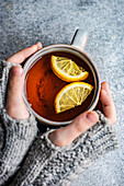 Kid hands in mittens hold cup of tea with lemon on grey concrete background