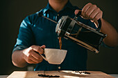 Crop anonymous male in casual clothes pouring freshly brewed coffee from French press pot into white ceramic cup standing at wooden table in kitchen