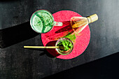 Top view of healthy matcha tea and dried powder near wooden chasen and chashaku on bright round stand on black table at sunlight