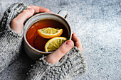 Kid hands in mittens hold cup of tea with lemon on grey concrete background