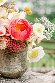 Shabby stone vase with bouquet of fresh peony and poppy flowers placed on terrace border on blurred background of lush trees in summer