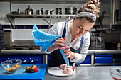 Female chef in apron using pastry bag to squeeze sweet cream on plate while cooking dessert on metal table in restaurant kitchen