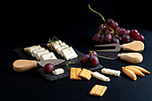 Top view of assorted delicious cheese arranged on black board with ripe grapes and crackers placed near knifes on dark surface