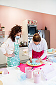 Focused female in masks and aprons standing at table while cooking delicious cake with blue glaze together