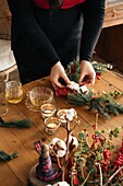 Hochformatiger Ausschnitt einer unkenntlichen Frau, die einen kreativen Weihnachtskranz aus dekorativen Tannen und roten Bändern herstellt, während sie zu Hause an einem Holztisch steht