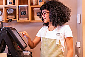 Young African American female seller in apron and glasses touching screen of modern cash register during work in zero waste grocery store