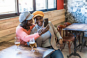 African American friendly females taking self shot on smartphone while sitting at table with glasses of beer in bar and enjoying weekend