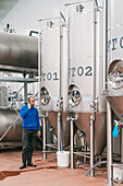Side view of male entrepreneur talking on cellphone against stainless steel vessels on wet floor in beer factory