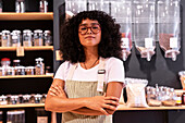 Self assured young African American woman in uniform and glasses with curly hair crossing arms and looking at camera during work in zero waste grocery shop