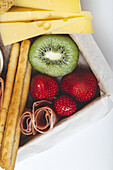 From above closeup brunch box with assorted sliced meats various types of cheese and crispbreads arranged near ripe cup kiwi sweet strawberries and peeled mandarin near jam in glass jar on colorful background