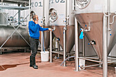 Side view of male entrepreneur talking on cellphone against stainless steel vessels on wet floor in beer factory
