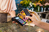 Crop unrecognizable client making contactless payment with mobile phone on POS terminal in floral shop