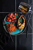 Lachssteak von oben mit würzigen Karotten und Kirschtomaten, serviert mit einem Glas trockenen Weißwein auf einem schwarz gekachelten Tisch
