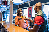 Cheerful African American couple clinking glasses with cold beer and proposing toast at counter in bar