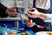 Crop female chef in blue apron taking bowl from male colleague while cooking dessert from crumbs and ground strawberries in restaurant kitchen