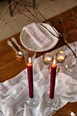 From above of white tablecloth and plates placed on festive table decorated with burning candles and dry branches of tree