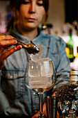 Crop female barkeeper putting ice cube with tongs into wineglass while preparing cocktail in bar with bottles on blurred background