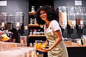Glad young African American woman in striped apron and glasses looking at camera with smile and putting glass box of dried mango on shelf during work in zero waste grocery store