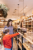 Positive mother with brown hair in dress standing with son in casual clothes pointing on glass jar with nuts placed on shelves in eco friendly store