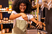 Soft focus of cheerful African American woman with curly hair using scoop to pack herbal tea from metal box into paper bag during work in eco friendly grocery shop