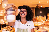 Happy young ethnic woman in uniform and glasses smiling and offering glass jar with nuts and diverse flours to camera while working in zero waste grocery shop