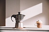 Geysir-Kaffeemaschine aus Metall auf dem Tisch mit aromatischem, schaumigem Latte Macchiato im irischen Glas in der Küche vor einer weißen Wand an einem sonnigen Tag