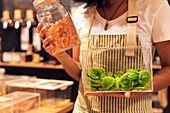 Cropped unrecognizable woman in uniform and glasses showing containers with dried fruits during work in zero waste grocery store