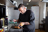 Focused bearded male putting pickled salted fish on toast served on board while standing at counter in kitchen of restaurant