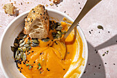 From above of spoon in white ceramic bowl with eaten pumpkin cream soup placed on table near scattered seeds and crispy bread pieces